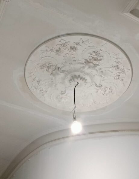 Plafond blanc avec un médaillon circulaire ornemental en stuc fleuri, un chantier de rénovation, et une ampoule LED simple suspendue par un fil noir.