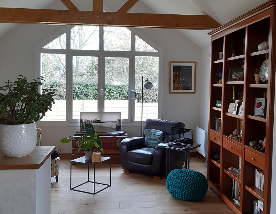 Salon lumineux avec grande fenêtre à pignon, fauteuil en cuir marron, bibliothèque en bois et plantes vertes, décor cosy.